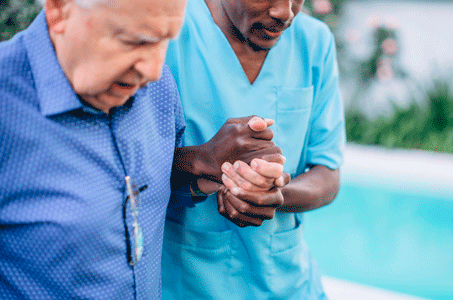 A nurse assists and old man by holding his hands.
